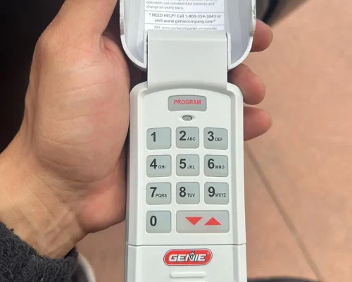 Garage Door Repair Huntington Beach 31 A hand holds a Genie garage door keypad with the cover flipped open, revealing a numeric keypad and “PROGRAM” button at the top. The Genie logo is visible near the bottom—perfect for those seeking reliable garage door repair Orange County services.